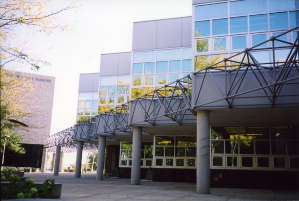 A kodak photo of a university art building on a sunny autumn day, taken from the courtyard near an entrance. There is a row of pillars and railing beneath the upper floor windows, creating a diagonal perspective in the frame, lower left in the background ramping up to the upper right in the foreground. A blue sky and yellow leaves are reflected in the upper floor windows. Those trees are just barely visible in the left of the frame. I think the architecture of this building could be described as postmodernism.