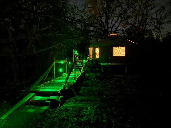 A night view of a bridge leading to a yurt. The bridge is lit with sparse green lights (LEDs) projecting circles of light on the bridge deck. The glow of green lights reflect onto ground around the bridge. The windows of the yurt and its plexiglass dome, glow with the warm light of comfort. The sky in the background is dark a few stars show between the leafless branches of an oak tree behind the yurt.