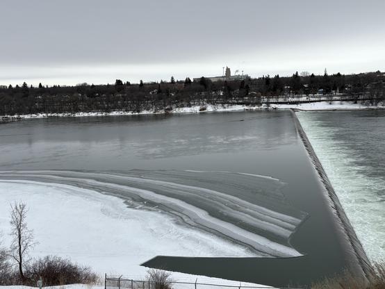 View over a wide partially frozen river with a small weir on the right. The ice on the left has a lovely striped pattern. A large chunk about the size and shape of a plane wing has broken off the ice shelf. On the other side of the ball one can see trees and some buildings. It’s a colour picture but the sky is overcast so everything is grey