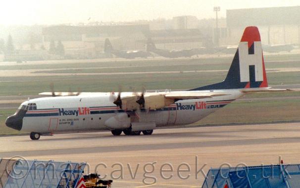 Side view of a grey, high-winged, 4 propellor-engined cargo plane taxiing from right to left.
The sides and belly are grey, while the top section is white, with a dark blue and red strip running along the side of the fuselage, overlaid with blue and red "HeavyLift" titles on both the front and rear of the fuselage.
The tail is red with a large, white uppercase letter "H" with a heavy, blue drop shadow.
Grey concrete apron fills the foreground, with blue metal frames on either side at the bottom, while large grassed areas divided by a runway lead up to cream coloured hangars with large military transport parked on the ramp in front, slowly vanishing into grey haze.