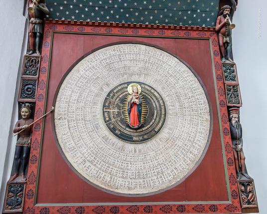 A large, rotating circular dial covered in concentric rings of writing, forming an astronomical and Saint's day calendar. At the centre is a statue of the Virgin Mary holding a baby Jesus.