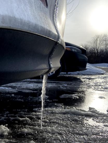 An icicle hanging from the bumper of a very dirty white car. There's another car parked next to it, and the bright yet hazy sun is reflecting in the icy slushy parking lot.