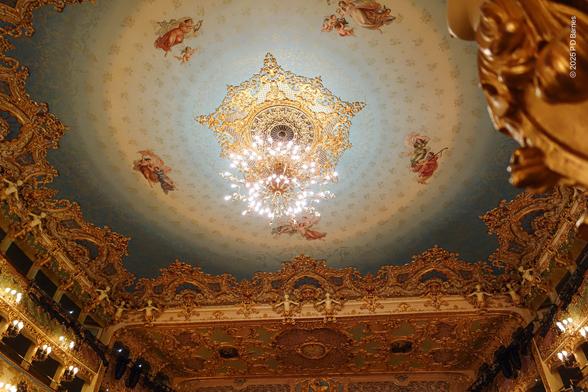 A giant chandelier in the ceiling of La Fenice opera house, Venice. It has an elaborate golden base, surrounded by radial patterns and paintings of the Graces, Music, Dance and Aurora. Surrounding that is heavily rococo gilt ornamentation featuring 22 nereids.