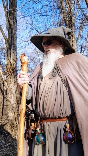 Middle aged man with grey beard, sunglasses, and a Gandalf style outfit with hat & staff looking off to the left. There is an array of colored spell bottles hanging along the belt.