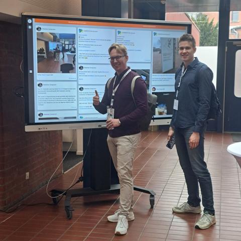 Blick auf einen großen Bildschirm mit Tröts zum Hashtag CLC25 
Davor stehen 2 Menschen und grinsen.
Weil sie auf der Wand mit dem Foto vorher und passendem Tröt erschienen sind