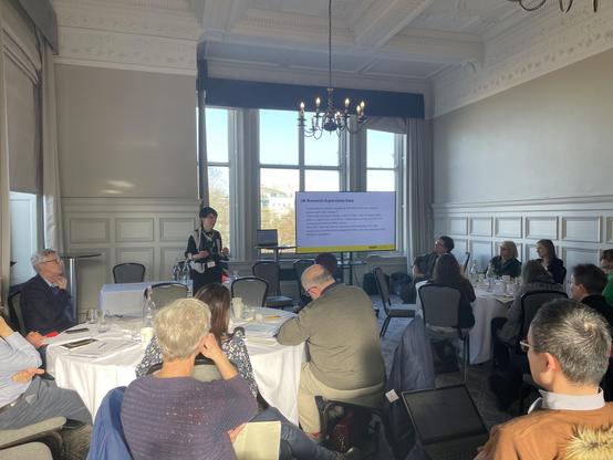 A speaker presents in a well-lit meeting room, surrounded by attendees seated at a round table. A projector displays information about UK Research Supervision Data. Attendees appear engaged, some taking notes. The room features large windows and decorative ceiling details.