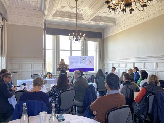 A presentation is taking place in a well-lit room with large windows. A speaker stands in front of a screen displaying information about addressing harassment and sexual misconduct, while an audience of around 20 people watches attentively, seated at tables.