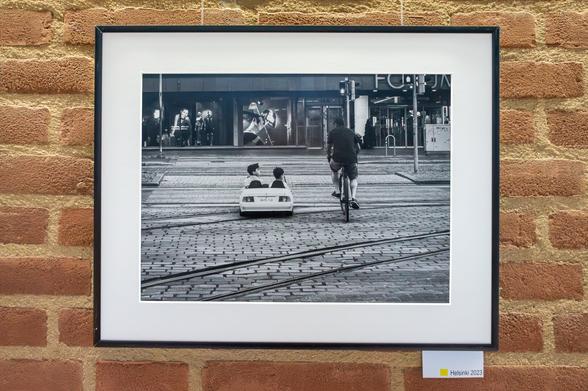 Foto einer Aufnahme, auf der zwei Kinder in einem Spielzeugauto sitzend, einen Mann auf dem Fahrrad neben ihnen ansehen.
