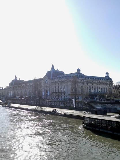 Das Musée d'Orsay in Paris , von der Passerelle Léopold Sédar Senghor gesehen: grosses Bahnhofsgebäude vom Anfang des 20. Jahrhunderts mit zwei immensen Uhren, direkt am Ufer der Seine.