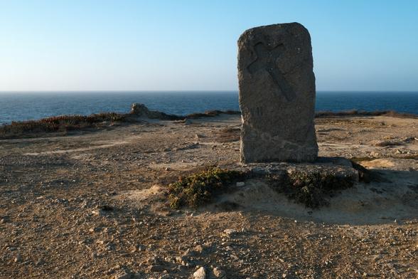 Photo of a stone, with a cross on it, placed on the top of a cliff. In the background the sea.