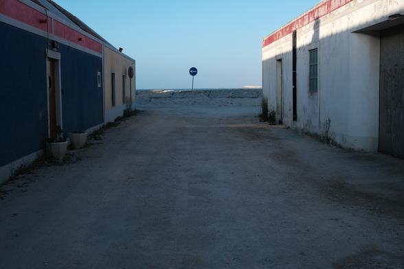 Photo of a small alley, with old, colored houses on both sides. At the end of the alley there is a traffic sign pointing to the left.