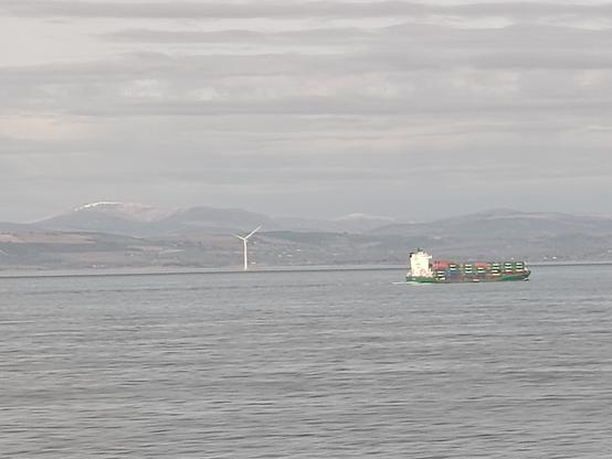 Picture of a cargo ship sailing past a wind turbine. In the background, a shoreline is visible, and in the far distance, mountains rise with late winter, snow topped, ridges.