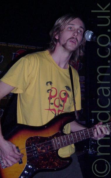 A man with long blond hair and a wispy beard, wearing a yellow t-shirt with black eyes in a v-shape overlaid by red swirls, playing a 4-stringed bass guitar.
He is in a dark room with black walls, and standing beside a microphone on a stand.
There is a green poster on the wall behind him advertising a New Years Eve party.