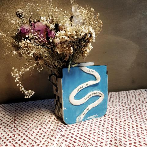 Ceramic slab built vase with poem on a banner across it. Light blue and black patterns decorate it. Styled with dried flowers against a gold wall. Made by Christine Lykkeh