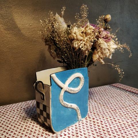 Ceramic slab built vase with poem on a banner across it. Light blue and black patterns decorate it. Styled with dried flowers against a gold wall. Made by Christine Lykkeh