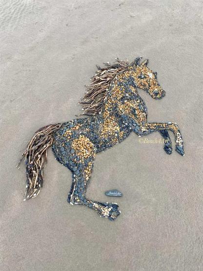 A completed horse artwork on Sandymere Beach, North Devon, UK, created with stones, shells, and driftwood. The detailed equine figure appears to be leaping, its mane flowing with driftwood strands, capturing a sense of movement and grace.