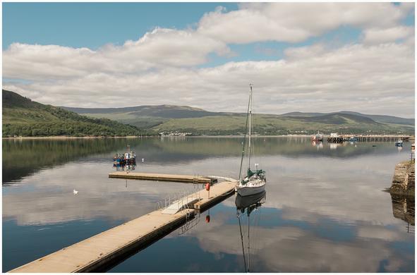 A tranquil harbor scene with a sailboat anchored near a wooden dock. Reflections of clouds and hills are visible on the calm water, surrounded by lush greenery and distant hills under a partly cloudy sky.