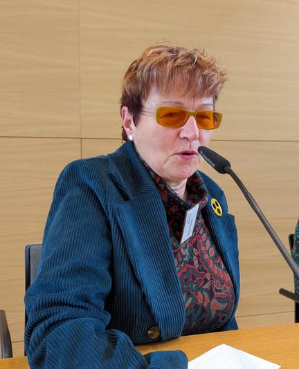 Nora Rämer spricht in ein Konferenzmikrofon, sie trägt eine Plakette mit drei schwarzen Punkten auf gelbem Grund als Zeichen dass sie blind ist.