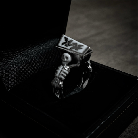A small silver ring inside an open black jewellery box. The ring consists of two skeletons facing away from each other and holding a coffin in between them, the skeletons and the coffin are decorated with white enamel.