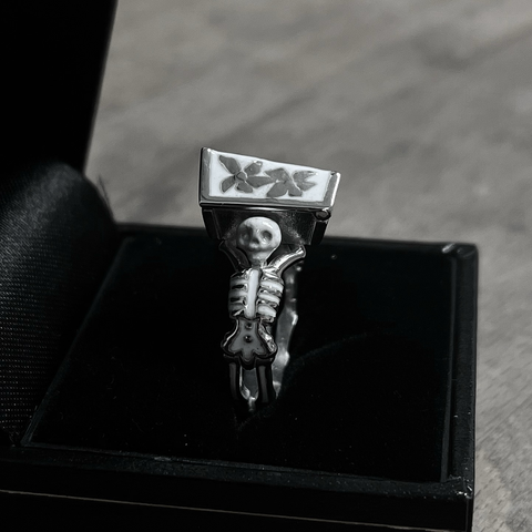 A small silver ring inside an open black jewellery box. The ring consists of two skeletons facing away from each other and holding a coffin in between them, the skeletons and the coffin are decorated with white enamel.