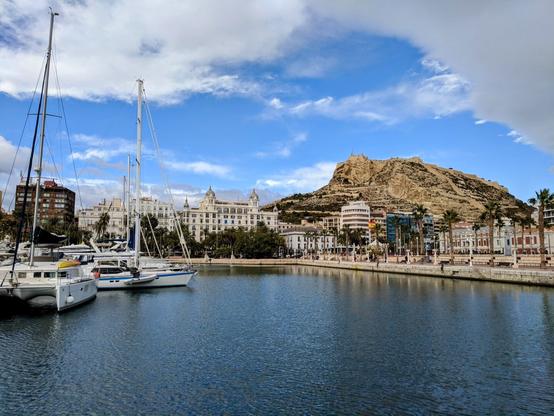 A harbour with sailing boats in the foreground, the city of Alicante in the midground, and a large hill, Mount Benacantil, with a castle on top in the background, under a partly cloudy sky.