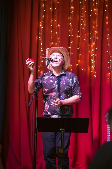 a man in a hat holding a maraca, singing into a microphone as he looks upwards towards us.  Behind him is a red curtain with some small yellow lights.