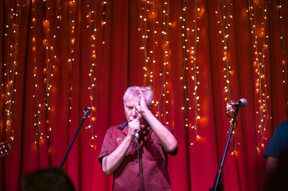 A man with grey hair and a dark red shirt sings into a microphone, holding his other hand up in front of his head. Behind him is a red curtain with some small yellow lights.