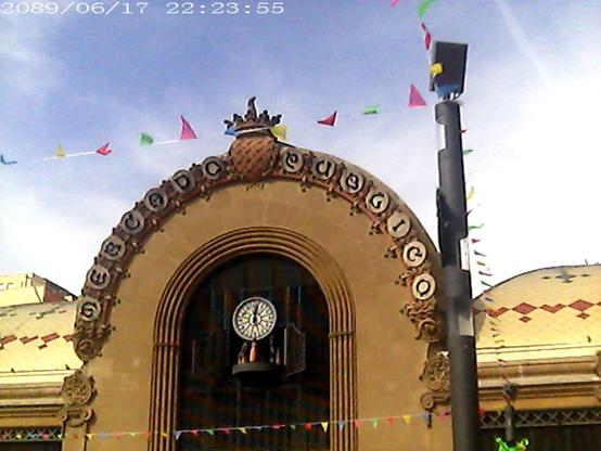 Clock and carrillon in a market building, small colored flags attached to a pole. The market building writes "Mercado Público" in a fancy handwriting font.
