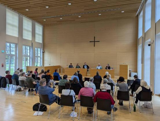 Ansicht des Podiums aus Blickrichtung der Zuhörerschaft. Pfarrer und Kirchenrat Gerald Kick sitzt vorn in der Mitte.