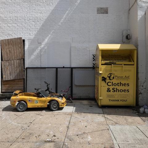 Yellow car with a 3 painted on the side for small children to die in on the sidewalk next to a yellow bin for donating old clothes