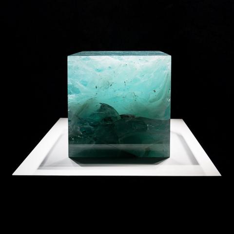Cube of of blue-green glass on a white pedestal in front of a black background