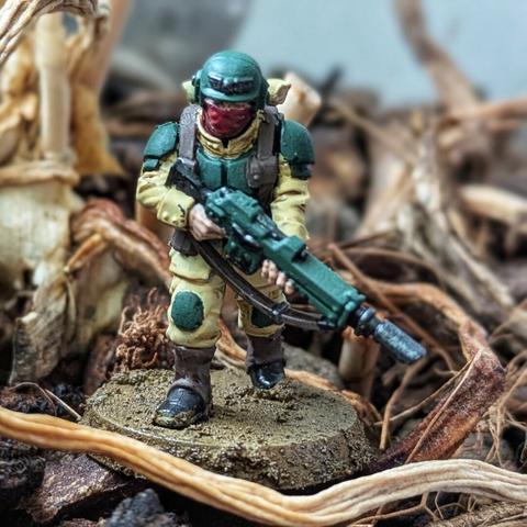 Figurka żołnierza Cadian 8th przedstawiona frontalnie, w dynamicznej pozie z lasgunem wycelowanym przed siebie. Zielony hełm i naramienniki harmonizują z beżowym mundurem oraz brązowymi butami. Na piersi widoczna torba w odcieniach brązu, a na plecach beżowy plecak. Podstawka wykonana w stylu bitewnym imituje ziemię pokrytą drobnymi kamieniami i otoczoną wysuszonymi roślinami, podkreślając atmosferę pola walki.