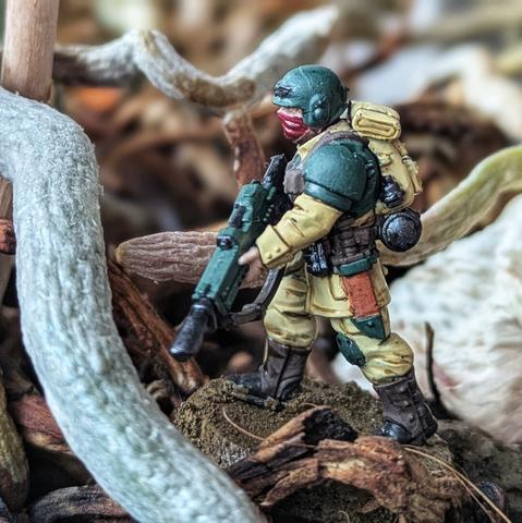 Figurka żołnierza Cadian 8th z uniwersum Warhammer 40K, widziana z boku. Żołnierz ubrany w klasyczny mundur Astra Militarum: beżowe spodnie i kurtkę z zielonymi naramiennikami oraz hełmem w tym samym odcieniu. Na plecach widoczny jasnobeżowy plecak oraz manierka w metalicznym kolorze. Detale, takie jak brązowe kabury i pas, podkreślają realistyczny charakter modelu. Podstawka stylizowana na ziemię, otoczona wysuszonymi korzeniami i gałęziami, tworzy surowy, naturalny klimat.