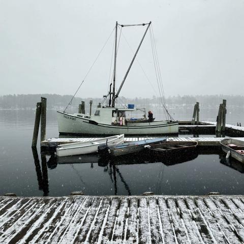 Looking out on a wooden dock where a lobster boat is moored. A fisherman is working on the back of the boat, and there is a light snow covering the dock. It's snowing and the water is still.