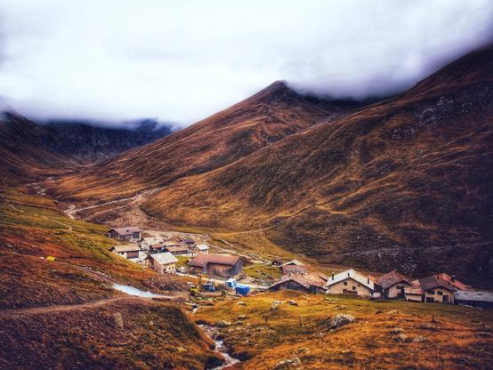This image captures the remote alpine village of Juf, located in the Avers Valley, Switzerland. Situated at an altitude of approximately 2,126 meters (6,975 feet), Juf is one of the highest permanently inhabited villages in Europe. The village consists of traditional stone and wood houses with sloped roofs, adapted to withstand the harsh mountain climate. Nestled among rolling hills and rugged peaks, Juf is surrounded by vast, treeless alpine meadows, characteristic of this elevation. A small stream winds through the valley, contributing to the headwaters of the Avers Rhine. The scene conveys the serene isolation of this unique high-altitude settlement, where life follows the rhythm of the mountains.
