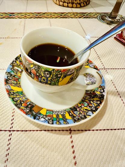 A cup and saucer for Turkish coffee creamers, served at a cafe in August. The china has a wide band encircling the surface, colorful mosaic art that appears modern yet primitive from North Africa. A cream woven table cloth is around it, with a spoon off the side inside the cup. There is a tiny colorful rainbow reflection in the center.