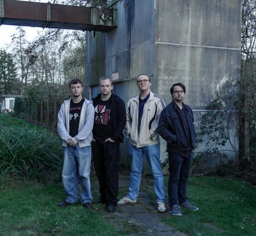 4 junge Männer in legeren Freizeitklamotten haben sich zu einem Gruppenbild zusammengestellt. Sie haben die Hände in die Taschen ihrer Jacken gesteckt und blicken entschlossen zur Kamera. Sie befinden sich draußen. Direkt hinter ihnen steht ein massiver Stahlbetonklotz mit einer Metalltür, aus dem nach links waagerecht ein rostiges Metallkonstrukt hinausragt. In weiterer Entfernung erkennt man Zäune und einen kleinen Bungalow.