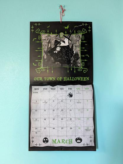 A wall calendar, open to March, and hanging on a blue wall. The calendar dates are listed on the bottom half on a white background with a thin cobweb patterns and a couple of Halloween theme icons in the form of a spider and a pumpkin head. The top half has a black border with cartoon-like luminous green framing in the form of gothic gates surrounding a central square with black, white, and grey artwork. The image is a broadly triangular character with black clothing, a rosette on his chest, a megaphone in his hand by his shouting mouth, and a tall, pointed, slightly bent hat on. Behind the character is the pale silhouette of a gothic nightmare town and some bats. Green writing below the image reads "Our town of Halloween."