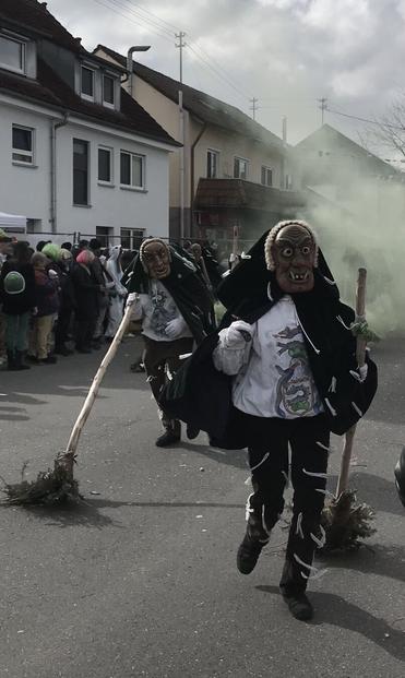Zwei schwäbische Narren mit hölzernen Masken auf einem Straßenumzzg, beide mit Reisigbesen, dahinter säumen Menschen die Straße, grünlicher Qualm steigt auf. Die Stimmung ist närrisch-dramatisch.