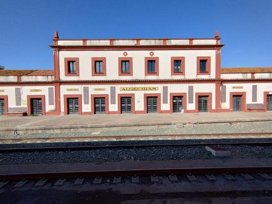 The old station building of Algeciras. Cream and terracotta fronting with Andalusian decorations against a blue sky. Marked on the station on yellow backgrounds above doors are the words Telegrago, Equipajes, Vestibulo, Algeciras, Jefe Estacion.