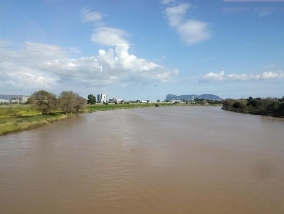 A brown river dominates the bottom half of a photo of Spain from a train. On the horizon, behind  some trees, can be seen Gibraltar. The sky is blue and there are some clouds.