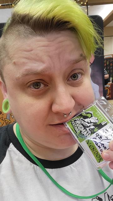 A greenhaired nerd in a white mothman baseball bites a name badge. "Forgman festival. Bug They/Them 9th Level Games"