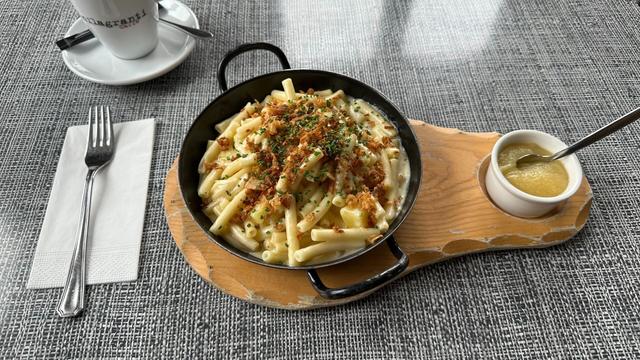 A single-serving size cast iron skillet of Älplermagronen (Swiss macaroni and cheese) on a wooden board served together with a small dish of apple sauce.