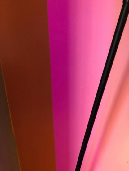Abstract image of a plain wall lit with pink light. On the left of the frame is a dark gold vertical section, seen at an angle. On the right a thin black metal rod diagonally crosses the wall.