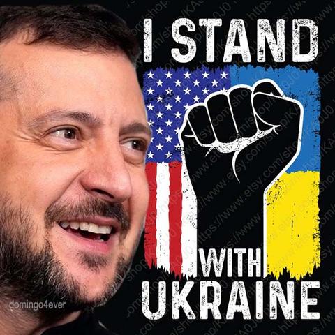 Hintergrund schwarz, links der Kopf von einem lächelnden Selenskyj. Rechts Hintergrund ein Streifen mit der Flagge der USA, daneben die Flagge der Ukraine darüber eine geballte schwarze Faust Weiß Text oben: I stand, weißer Text unten : with Ukraine.
