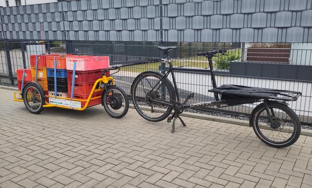Ein schwarzes Lastenrad ist mit einem großen dreirädrigen Fahrradanhänger verbunden. Der Anhänger ist mit vier Stapeln Bäckerkisten beladen, die hintereinander auf der Ladefläche verzurrt sind.