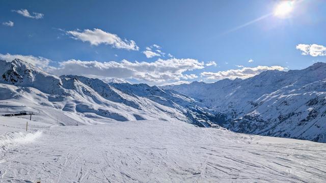 A panoramic winter landscape taken from the top of the Karbahn in Hochgurgl ski resort. The image shows a vast expanse of pristine snow-covered slopes with ski tracks visible in the foreground. Majestic snow-capped mountains of the Ötztal Alps extend across the horizon, creating a dramatic alpine valley. The scene is illuminated by bright sunlight under a vibrant blue sky dotted with wispy white clouds. Some ski infrastructure is visible on the left side of the image. This vantage point offers a spectacular high-altitude view of the surrounding alpine terrain, with the characteristic deep valley formation of the Ötztal region visible in the distance.