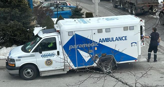 Photo of an ambulance parked on a street beside a house. A paramedic is sitting in the driver’s seat. Another paramedic is opening the door at the back of the ambulance.