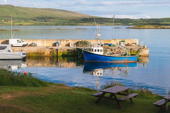 A scenic view of a harbour in Ahakista, Sheep's Head, Cork, Ireland, featuring fishing boats moored alongside a concrete pier, with a picnic table in the foreground.