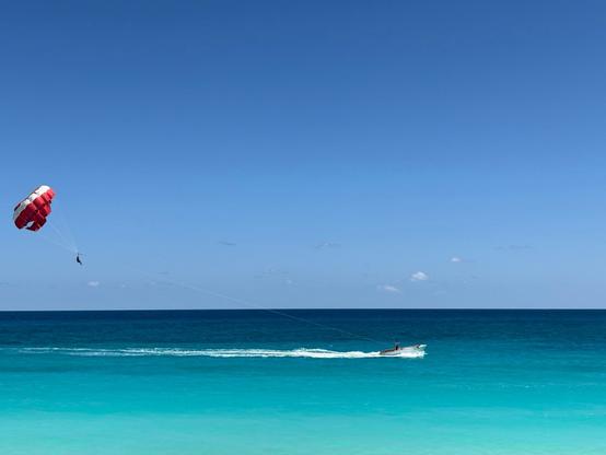 Parasailing activity over crystal-clear turquoise waters. A red and white parachute carries a person high in the air, connected by a long line to a small motorboat creating a white wake below. The scene features a brilliant blue sky with minimal clouds and a striking gradient of blue ocean colors from deep navy at the horizon to bright turquoise near the shore.​​​​​​​​​​​​​​​​